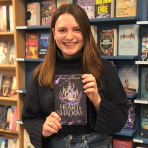 Unsere Buchhändlerin hält das Buch "Heart & Shadow" von Marie Graßhoff in der Hand, zu dem sie eine Rezension geschrieben hat.